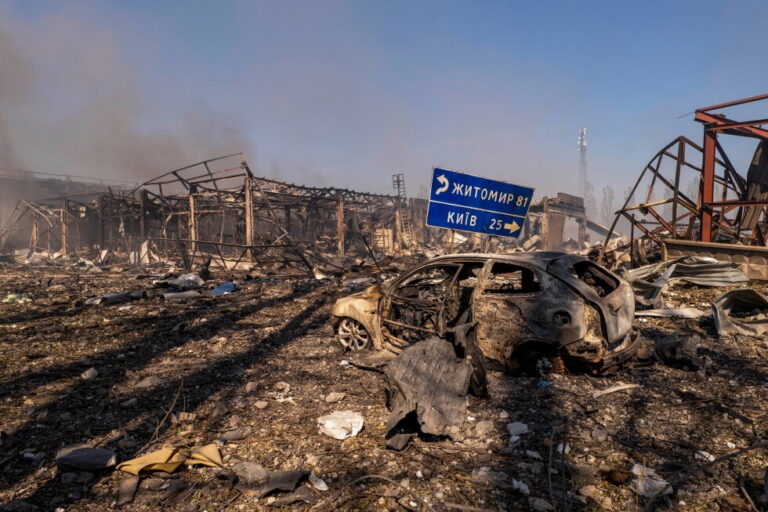 A view of a destroyed car amid damaged buildings, with a road sign showing the direction and distance to Kyiv and Zhytomyr
