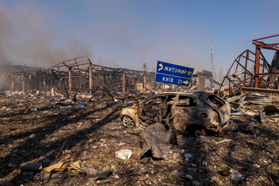A view of a destroyed car amid damaged buildings, with a road sign showing the direction and distance to Kyiv and Zhytomyr