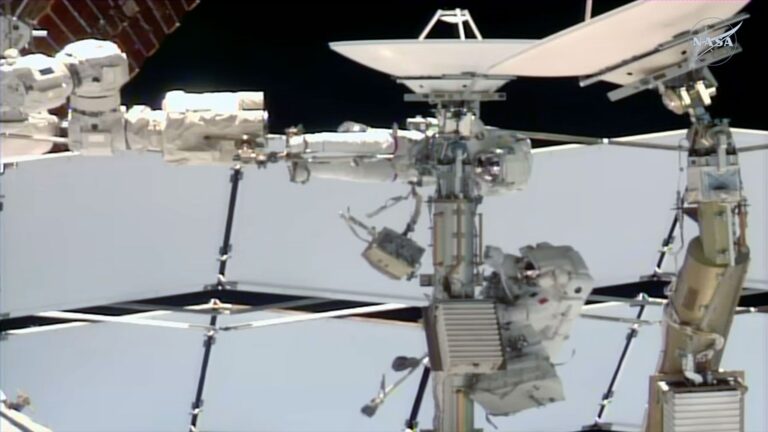 two astronauts dressed in white spacesuits work near a couple of antenna dishes outside of a space station
