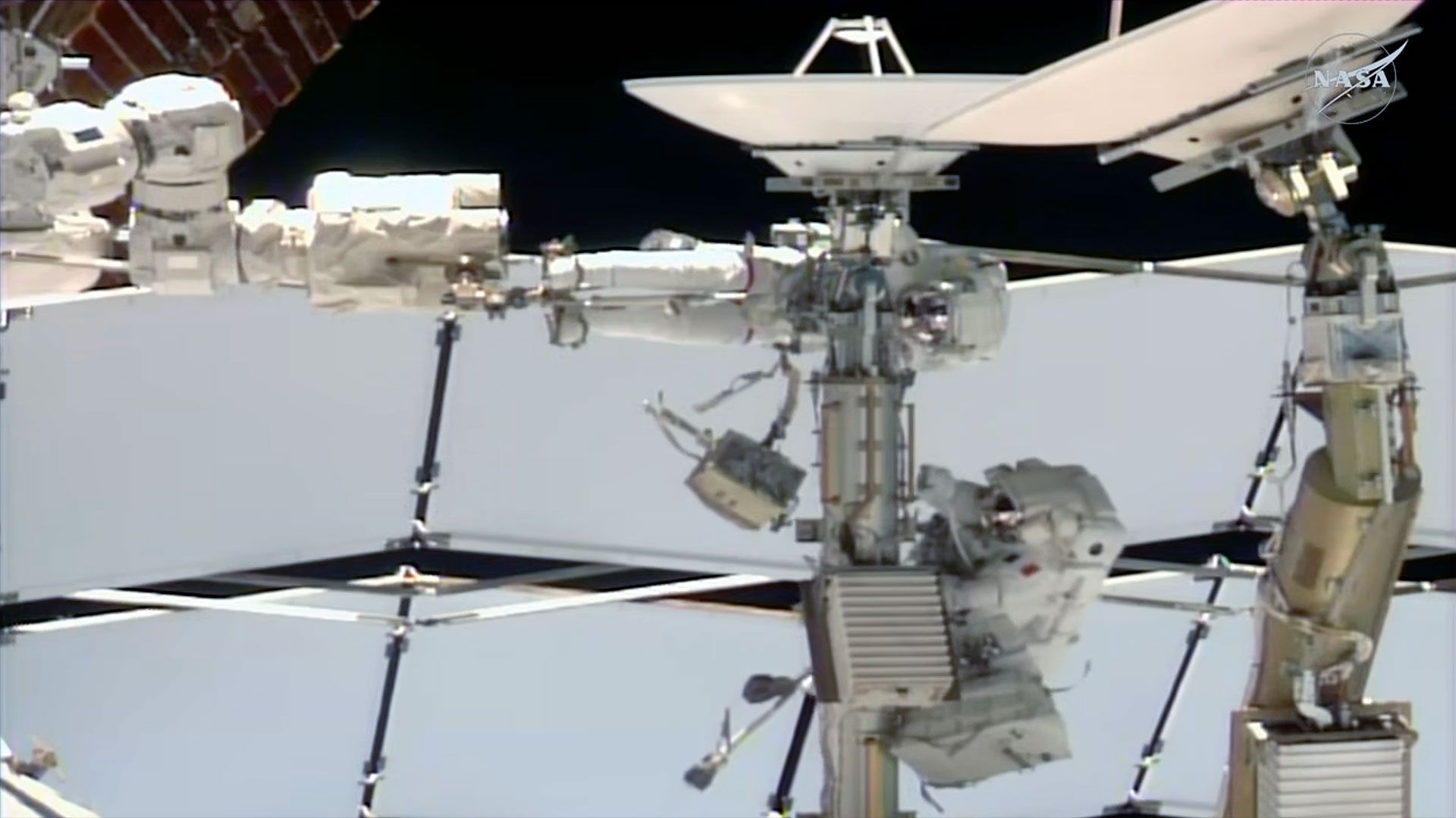 two astronauts dressed in white spacesuits work near a couple of antenna dishes outside of a space station