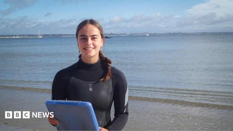 Kira Dunn smiles at the camera as she stands on a beach. She is wearing a wetsuit and holding a blue tablet