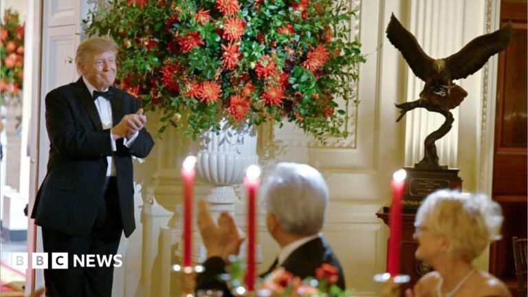 Trump, in a tuxedo, stands smiling next to a large white vase filled with red flowers. To the left is a large bronze statue