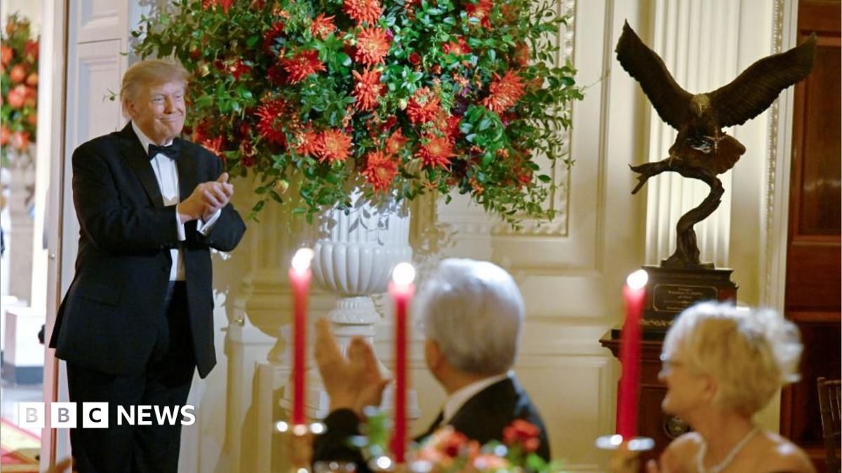 Trump, in a tuxedo, stands smiling next to a large white vase filled with red flowers. To the left is a large bronze statue