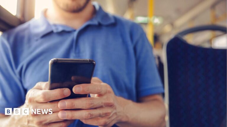 A man in a blue Tshirt holding a mobile phone and sitting on a bus