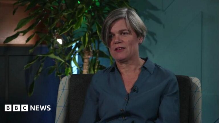 A middle-aged woman with shortish grey hair. She is wearing a turquoise shirt against the backdrop of an indoor plant