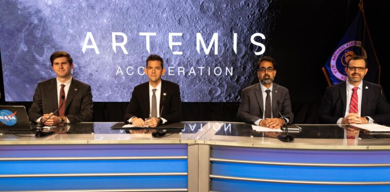 Four people sit behind a curved desk with microphones in a studio setting. A large screen behind them displays the words
