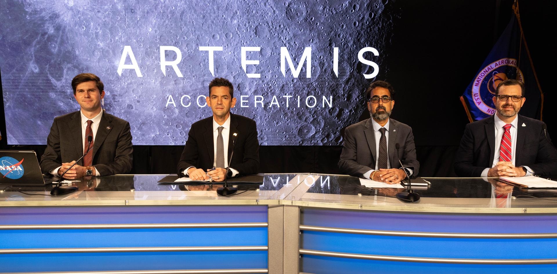 Four people sit behind a curved desk with microphones in a studio setting. A large screen behind them displays the words