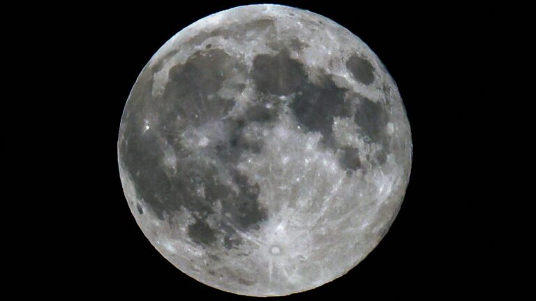 A photo of a full moon shining in a black sky, with dark lunar seas and bright craters visible on its surface