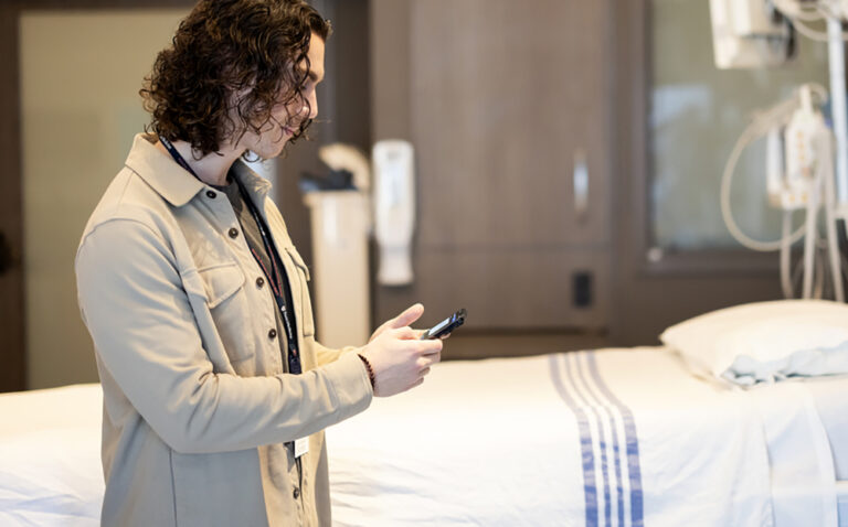A patient standing next to a hospital bed typing on their smartphone