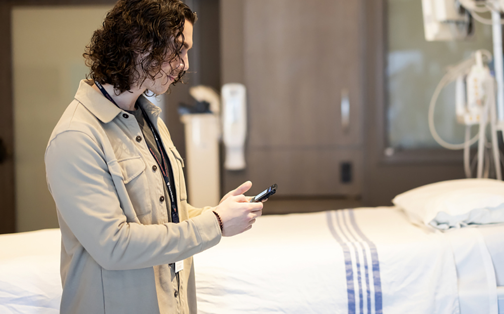A patient standing next to a hospital bed typing on their smartphone