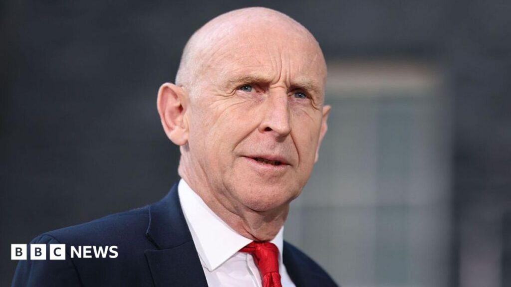 John Healey stands looking into the middle distance wearing a red tie, white shirt and navy blue suit. There is a blurred