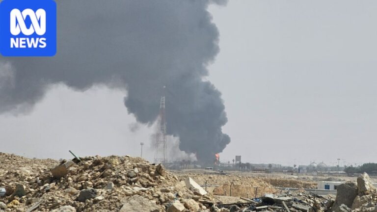 A large cloud of black smoke and flames rise out of an oil facility with rubble in the foreground