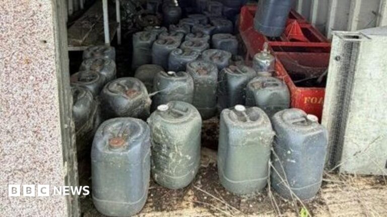 A number of dark green containers inside what appears to be an open building. There are two red crates on one side. The