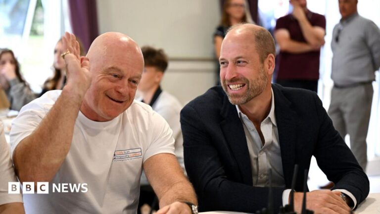 Ross Kemp waves one hand in his air as he smiles. He is sat next to Prince William who laughs as he looks at Kemp