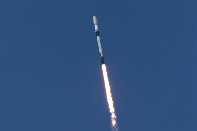 A white rocket with black bands ascends into a clear blue sky with a fiery plume of exhaust coming from the rocket