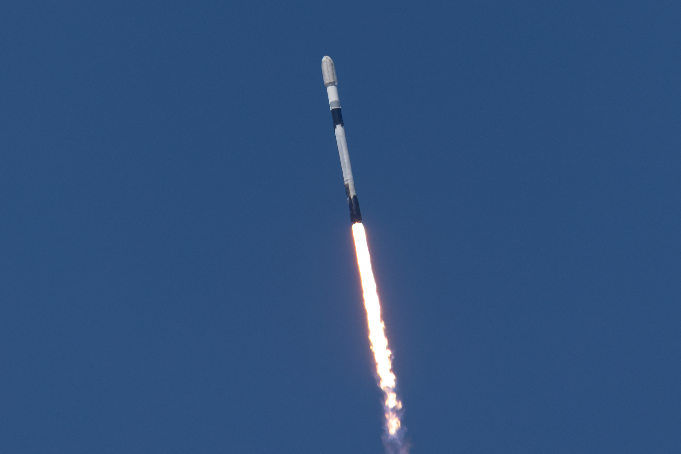 A white rocket with black bands ascends into a clear blue sky with a fiery plume of exhaust coming from the rocket
